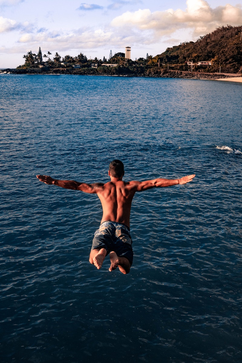 Back view of unrecognizable shirtless muscular male traveler jumping into blue sea water with outstretched arms on sunny day