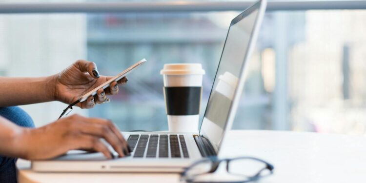 Close-up of hands using a laptop and phone with coffee on a modern office desk.