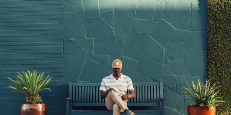 A man sitting on a bench with a green wall in an urban outdoor setting, reading his phone.