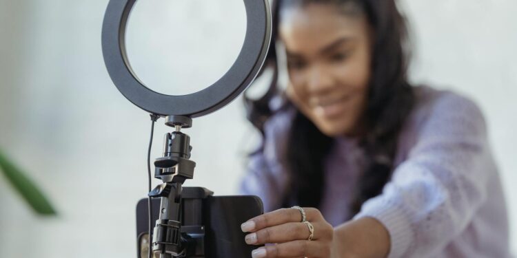 Cheerful young African American female blogger in stylish sweater smiling while setting up camera of smartphone attached to tripod with ring light before recording vlog