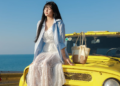 A stylish woman in a white dress sits on a yellow jeep, enjoying a sunny coastal day.