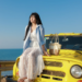 A stylish woman in a white dress sits on a yellow jeep, enjoying a sunny coastal day.