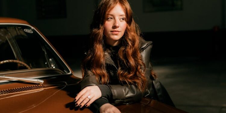 Portrait of a young woman leaning on a vintage car inside a dimly lit garage.