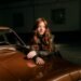Portrait of a young woman leaning on a vintage car inside a dimly lit garage.