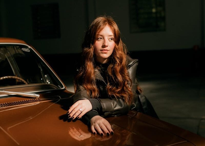 Portrait of a young woman leaning on a vintage car inside a dimly lit garage.