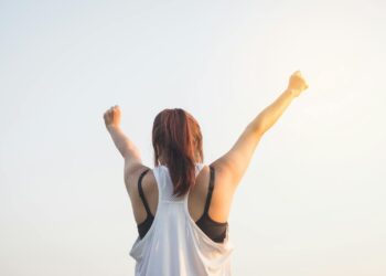 A woman celebrates success with arms raised in a bright outdoor setting.