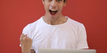 Young man in white shirt with laptop, celebrating success against a vibrant red background.