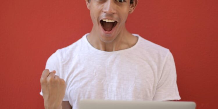 Young man in white shirt with laptop, celebrating success against a vibrant red background.
