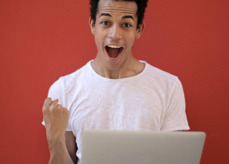 Young man in white shirt with laptop, celebrating success against a vibrant red background.