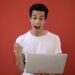 Young man in white shirt with laptop, celebrating success against a vibrant red background.