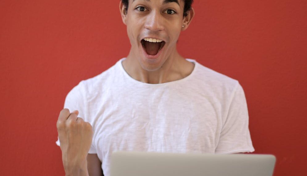 Young man in white shirt with laptop, celebrating success against a vibrant red background.