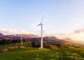 A wind turbine stands tall against a mountainous backdrop under a vibrant sky at sunset.