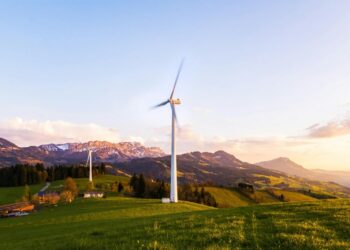 A wind turbine stands tall against a mountainous backdrop under a vibrant sky at sunset.