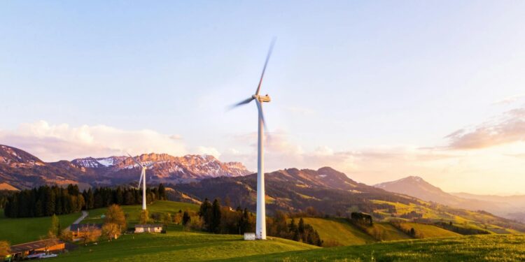 A wind turbine stands tall against a mountainous backdrop under a vibrant sky at sunset.