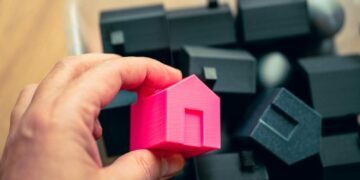 Hand selecting a pink house model from a box of miniature homes, symbolizing real estate choices.