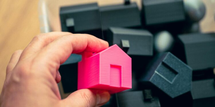Hand selecting a pink house model from a box of miniature homes, symbolizing real estate choices.