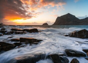 Vibrant sunset illuminates ocean waves crashing on a rocky shore.