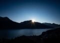 Sun rising behind dark mountains above a calm blue lake