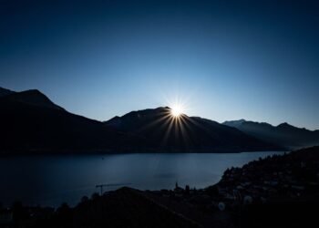 Sun rising behind dark mountains above a calm blue lake