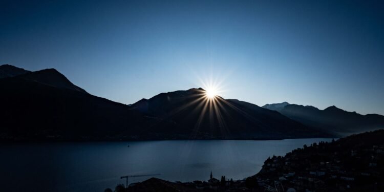Sun rising behind dark mountains above a calm blue lake