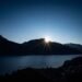 Sun rising behind dark mountains above a calm blue lake