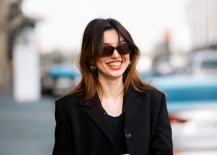 A woman in sunglasses and a black coat smiling on a city street, Positive Phil