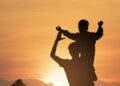 Silhouette of an adult carrying a child on their shoulders against a warm sky