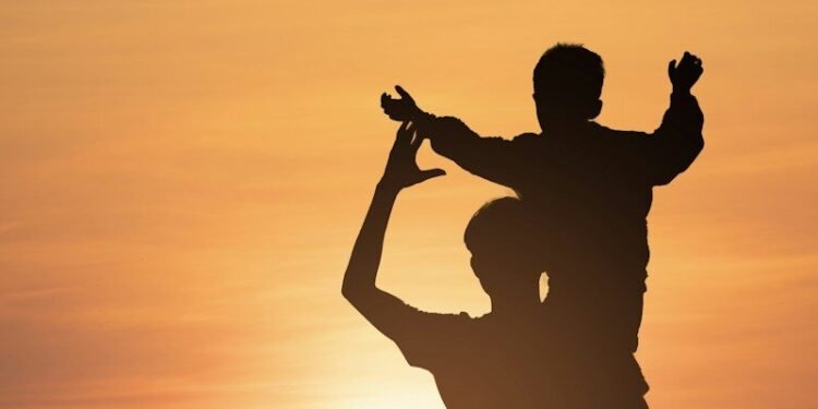 Silhouette of an adult carrying a child on their shoulders against a warm sky