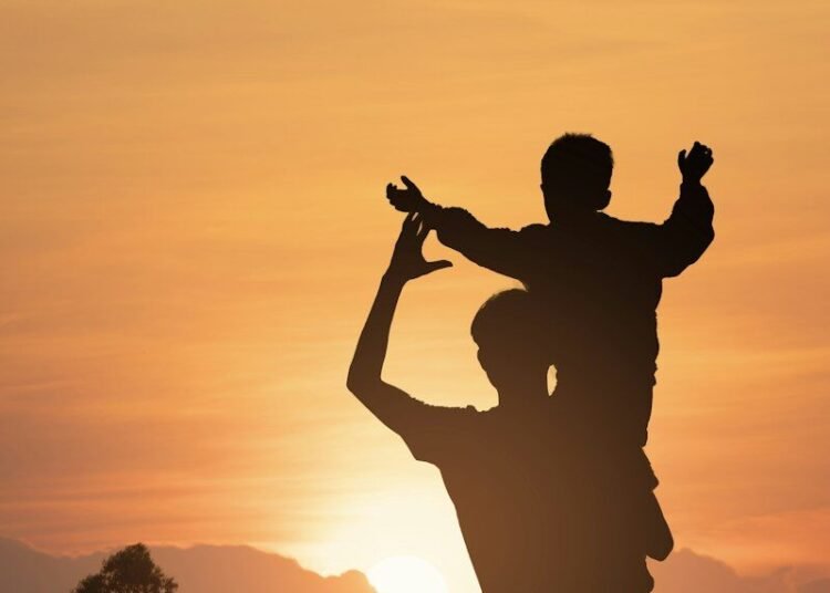 Silhouette of an adult carrying a child on their shoulders against a warm sky