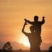 Silhouette of an adult carrying a child on their shoulders against a warm sky